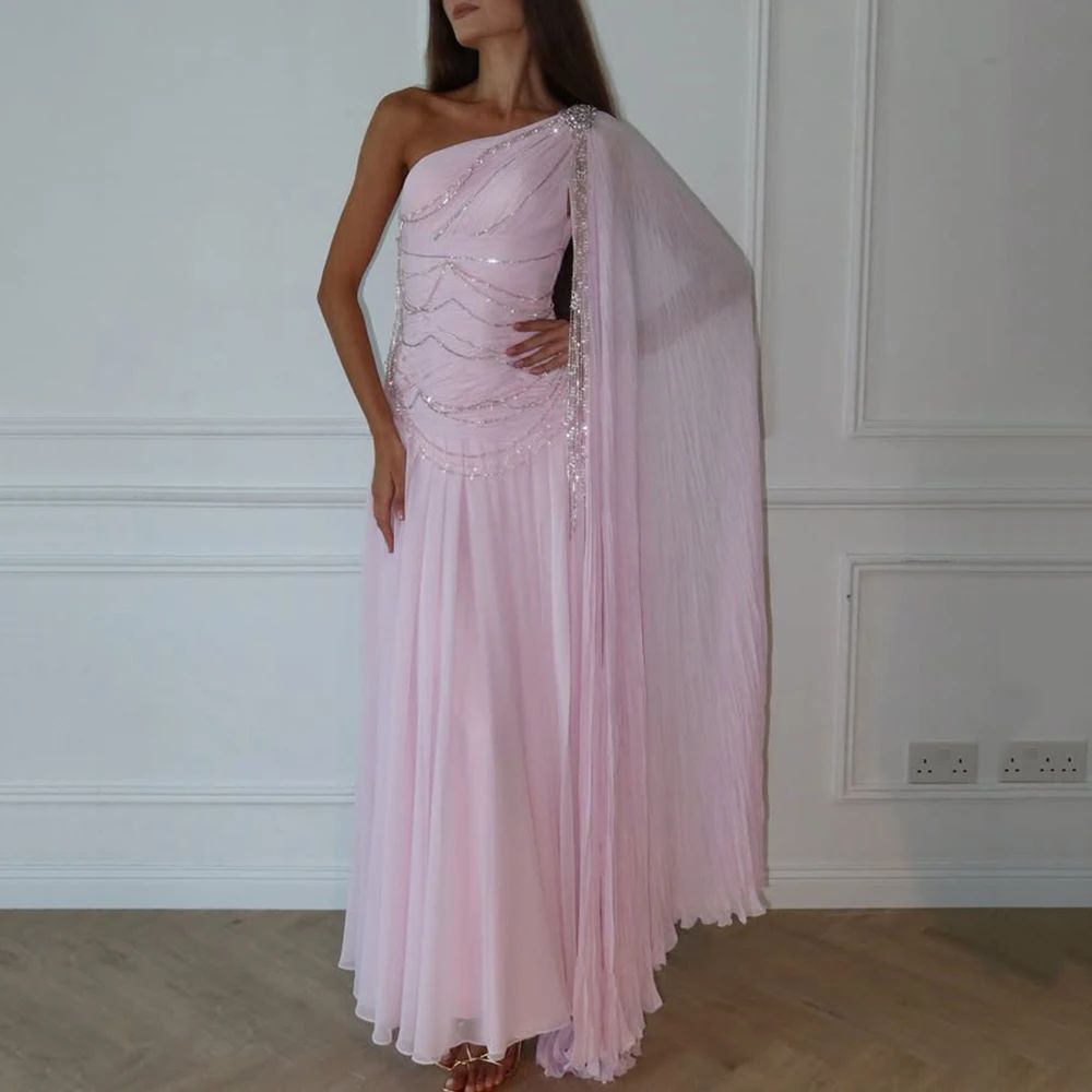 Woman wearing a pink one-shoulder dress with a cape against a white wall.
