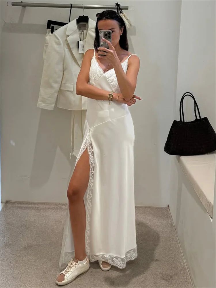 Woman taking a mirror selfie wearing a white dress with a high slit, standing in a room with a white wall and black handbag.