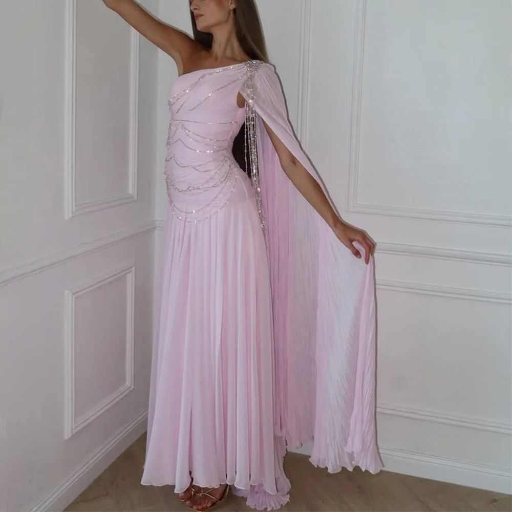 Woman wearing a pink evening gown with a sheer overlay against a white paneled wall.