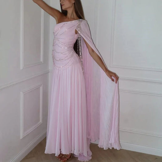 Woman wearing a pink evening gown with a sheer overlay against a white paneled wall.