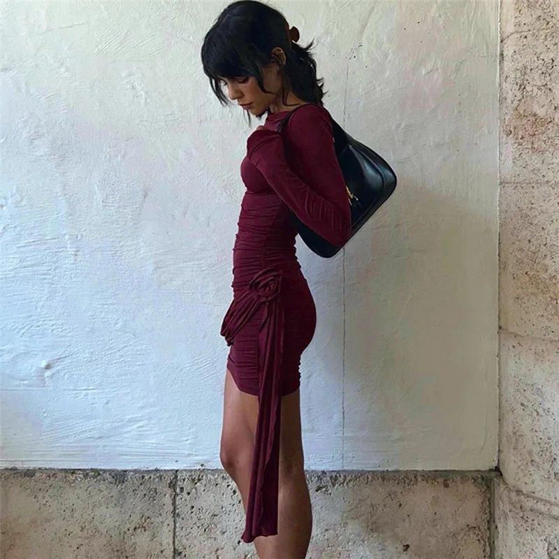 Woman in a burgundy dress holding a black handbag against a textured wall.